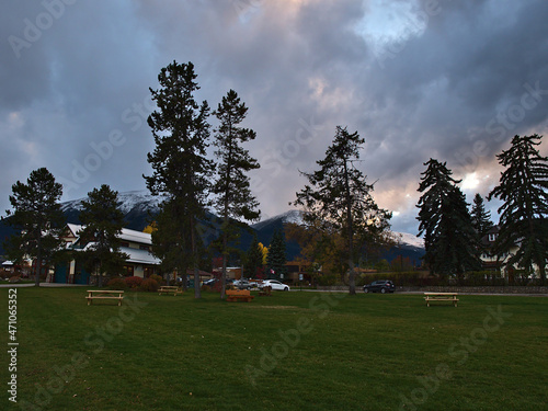 View of empty park area in small town Jasper, Alberta, Canada in the evening with green grass, benches and trees with snow-capped mountains. © Timon
