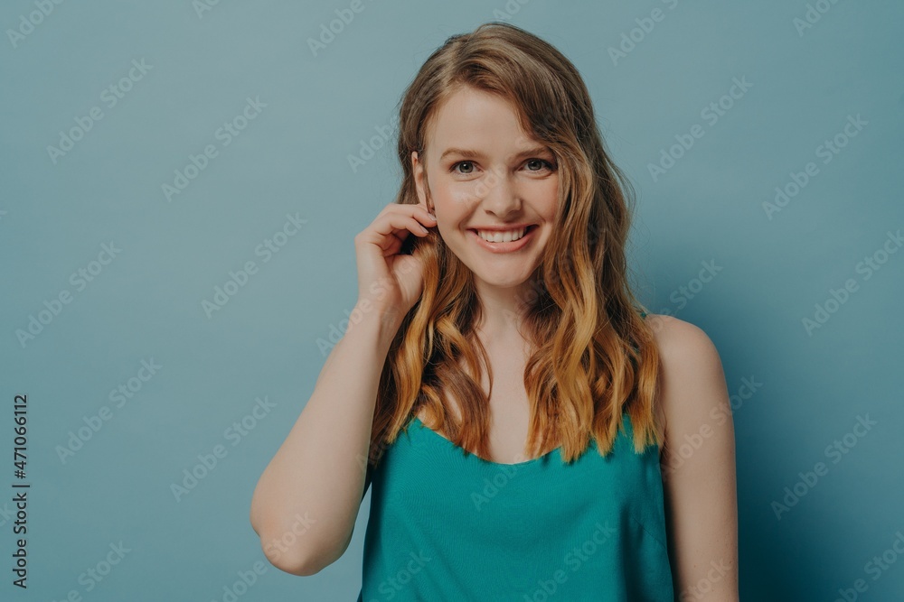 Charming female model looking at camera with beaming smile, tucking wavy styled hair behind her ear Charming female model looking at camera with beaming smile, tucking wavy styled hair behind her ear