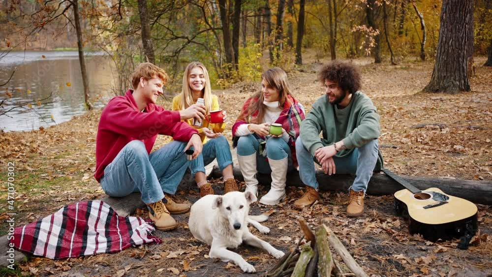 Young friends, men and women, smiling and enjoying hot tea from thermos, sitting on log in autumn forest near river. Slow motion Young friends, men and women, smiling and enjoying hot tea from thermos, sitting on log in autumn forest near river. Slow motion