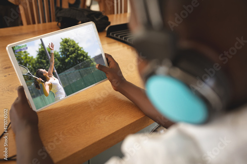 African american boy wearing headphones at home watching female tennis player on tablet
