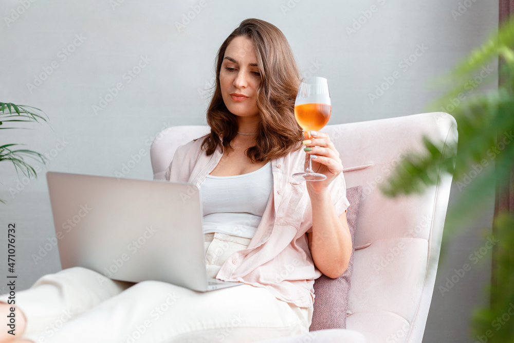 Young beautiful woman sitting on armchair drinking rose wine and using laptop. Caucasian relaxed woman watching movie on laptop or make online purchases with glass of wine. Young beautiful woman sitting on armchair drinking rose wine and using laptop. Caucasian relaxed woman watching movie on laptop or make online purchases with glass of wine.