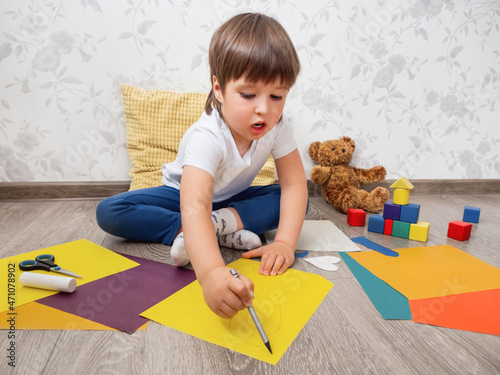 Toddler boy learns to cut colored paper with scissors. Kid sits on floor in kids room with toy blocks and teddy bear. Developing feeling sensations and fine motor skills at home. photo