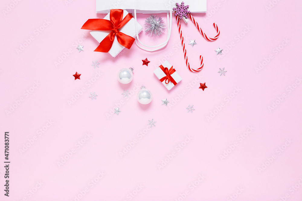 Fototapeta premium Christmas, New Year composition. White paper bag with presents, sweet candy canes, holiday decorations, silver balls, glitter confetti stars on pastel pink background. Flat lay, copy space.