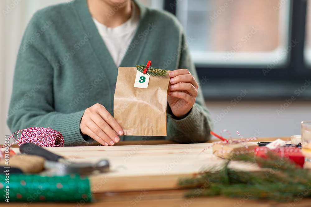 Obraz premium christmas, winter holidays and hobby concept - close up of woman with clothespin, craft paper bag and tag packing gift and making advent calendar at home