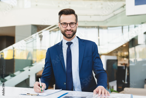 Successful caucasian smiling man shop assistant receptionist in formal attire writing looking at camera while standing at reception desk in hotel car dealer shop photo