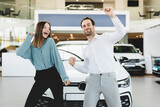 Excited dancing cheerful caucasian young family couple feeling happy after buying purchasing winning new expensive car auto at automobile dealer shop store