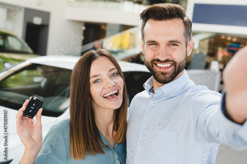 Selfie vlogging blogging on smart phone of happy young family holding car keys after buying new automobile at dealer shop store. Heterosexual husband and wife purchasing expensive auto