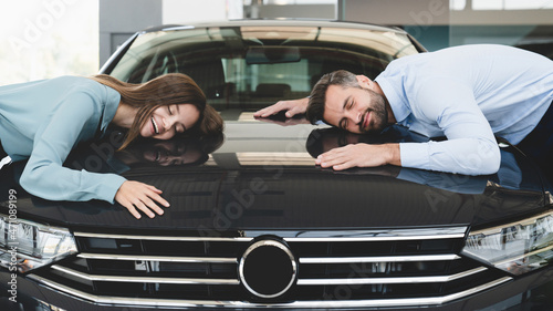 Closeup of extremely satisfied happy caucasian young couple family husband and wife hugging embracing their new car, feeling excited after buying expensive auto at dealer shop store.