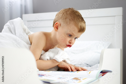 A little boy voices a book that he is looking at while lying in a crib under a blanket