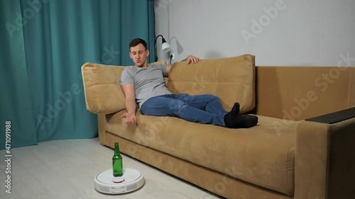 White robot vacuum cleaner delivers alcohol beer bottle to brunet short-haired man sitting on sofa in grey t-shirt, jeans in living room. photo