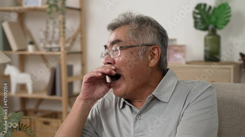 old asian man in glasses yawning before going asleep in bright living room at home. elderly male with grey hair feeling tired while sitting on couch. senior people cover opened mouth by hand on sofa