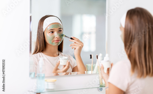 beauty  skin care and people concept - teenage girl with brush applying clay mask to face and looking in mirror at bathroom