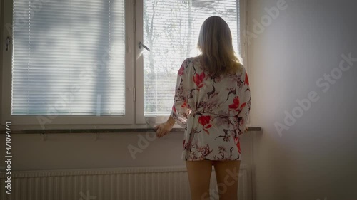 Beautiful young blonde with long hair by the window opens the blinds. Sunny rays light. In the morning slender girl in the sunlight looks out the window and enjoys her good positive mood.
