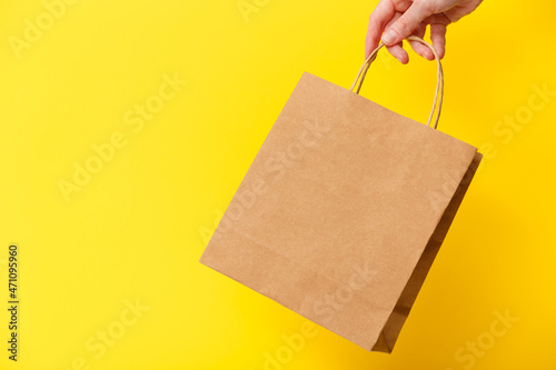 Female hand holding brown craft blank shopping bag isolated on yellow background. Black friday sale, discount, recycling, shopping and ecology concept.