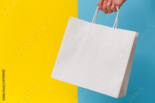 Female hand holding white blank shopping bag isolated on yellow and blue background. Black friday sale, discount, recycling, shopping and ecology concept.