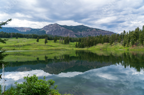 Trout Lake Trail in Yellowstone National Park on a cloudy summer day - a mountain reflects in a blue lake  with green grass on the hillside in the distance