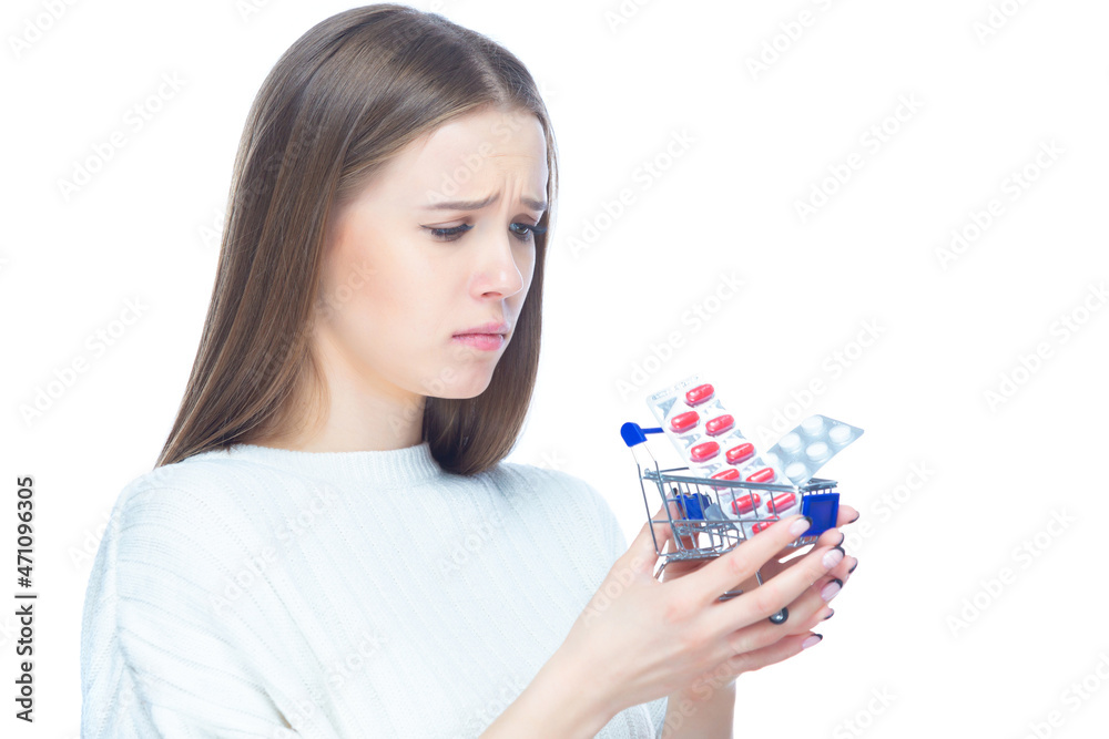 Studio shot of upset disappointed girl holding in her hands a small shopping cart full of expensive pills and medicine, isolated on white background Studio shot of upset disappointed girl holding in her hands a small shopping cart full of expensive pills and medicine, isolated on white background