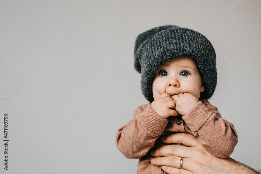Baby wearing a grey oversized knitted beanie, sucking fingers, on white wall background. Baby wearing a grey oversized knitted beanie, sucking fingers, on white wall background.