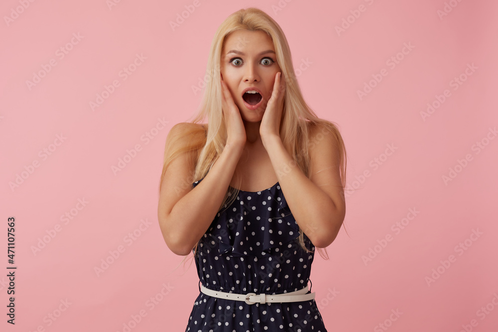 Studio Portrait of blonde female wears polka dot dress keeps mouth widely opened with surprised facial expression, touching her cheeks. isolated over pink background Studio Portrait of blonde female wears polka dot dress keeps mouth widely opened with surprised facial expression, touching her cheeks. isolated over pink background