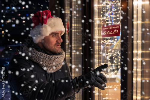 Man in Santas hat looking and dreaming in illuminated shop window. Christmas holidays sales concept