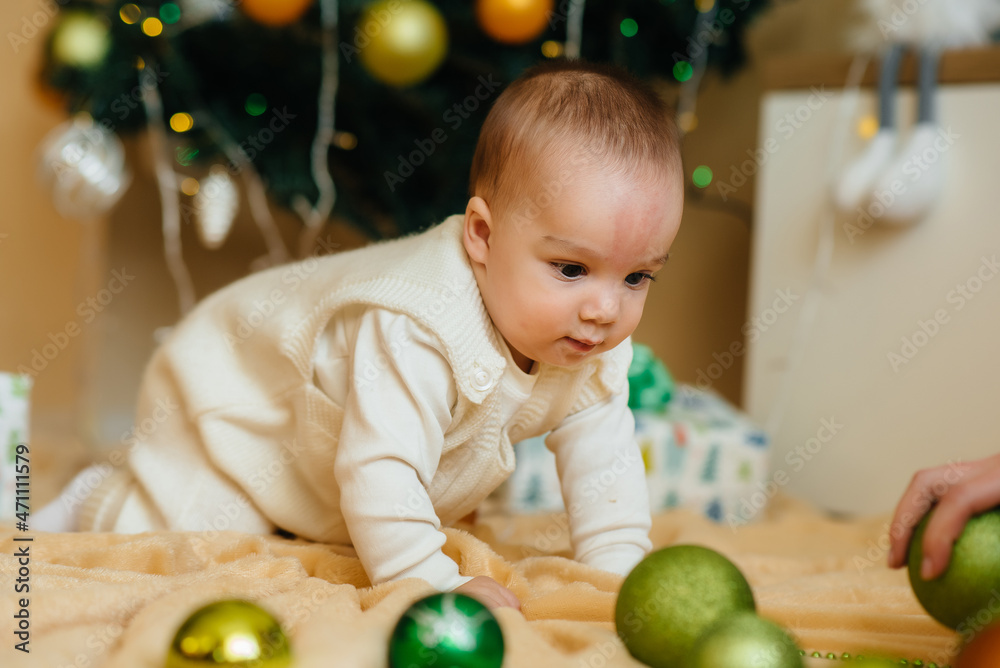 Fototapeta premium Cute smiling baby is lying under a festive Christmas tree and playing with gifts. Christmas and New Year celebrations