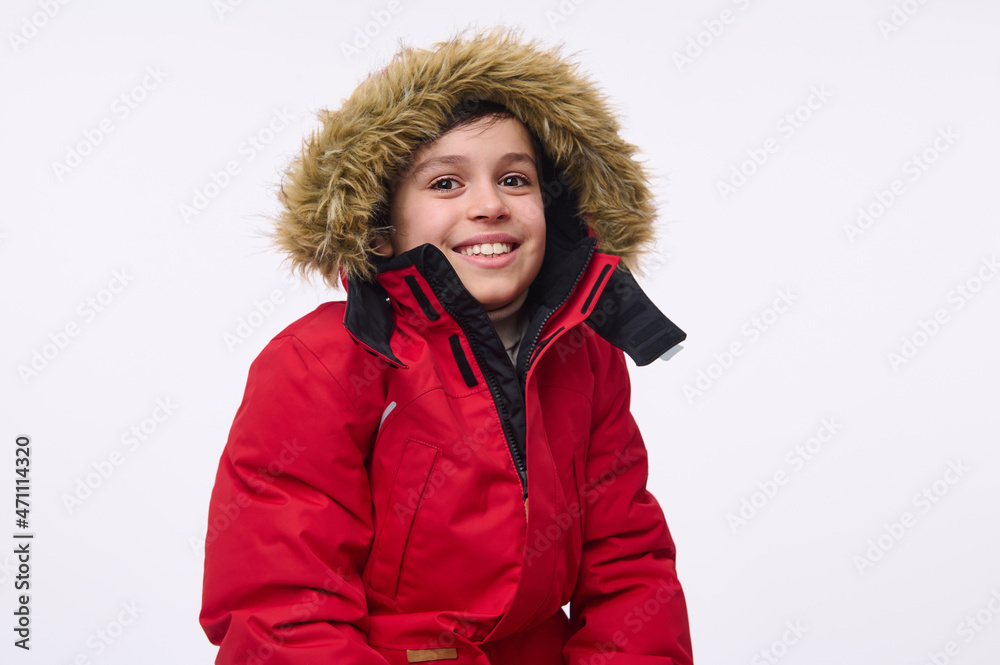 Waist length portrait of handsome cheerful preadolescent, school aged boy wearing bright red down jacket with hood, smiling toothy smile looking at camera isolated on white background with copy space Waist length portrait of handsome cheerful preadolescent, school aged boy wearing bright red down jacket with hood, smiling toothy smile looking at camera isolated on white background with copy space