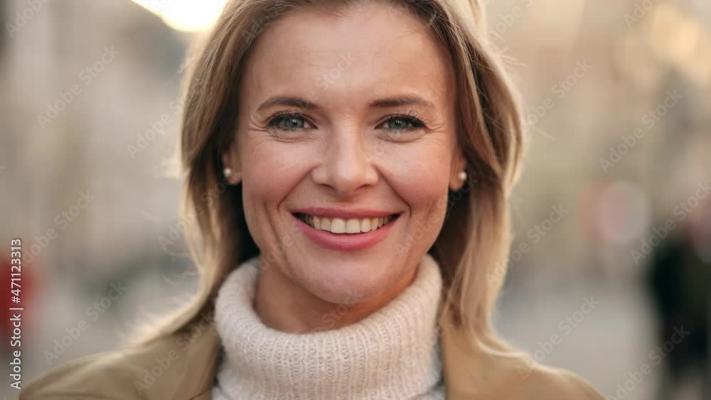 Lovely middle-aged blond woman with a beaming smile and looking at the camera. Happy adult woman smiling with teeth smile outdoors and stay on city street at sunset time Lovely middle-aged blond woman with a beaming smile and looking at the camera. Happy adult woman smiling with teeth smile outdoors and stay on city street at sunset time
