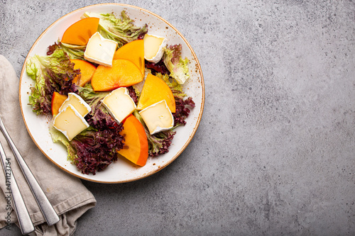 Top view flat lay persimmon salad with brie cheese  fresh salad leaves on white plate stone gray background  seasonal fruit salad as appetizer  vegetarian healthy food space for text