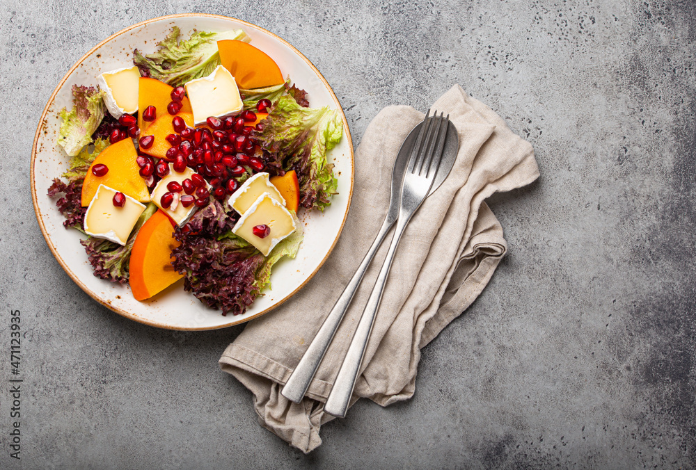 Top view flat lay persimmon salad with brie cheese, fresh salad leaves, pomegranate seeds on white plate stone gray background, seasonal fruit salad as appetizer, vegetarian healthy food copy space