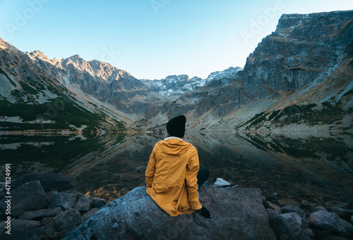 Woman in yellow jacket sitting relaxed on the shore of Czarny Staw Gąsienicowy lake, crystal clear blue water in the tatra mountains in poland. Tourist destination in europe.