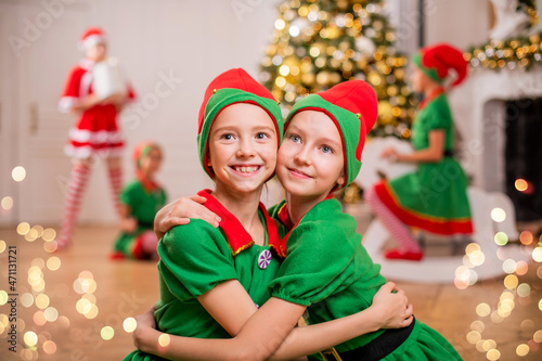Two cheerful happy girls-friends in costumes of Santa's elves on the background of a Christmas tree and other children