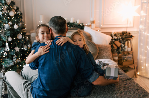 a happy father hugs two young daughters sitting on a bed near a Christmas tree. family happiness and joy