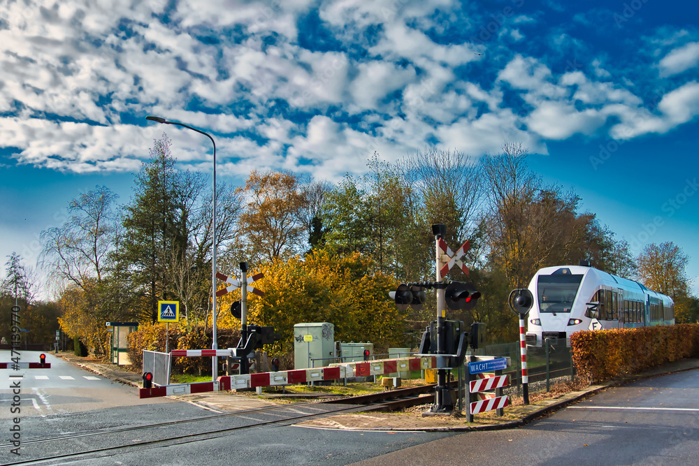 Local train approaching a railway crossing with red lights and barrier on a sunny autumn day. Baflo, province of Groningen, Netherlands.
