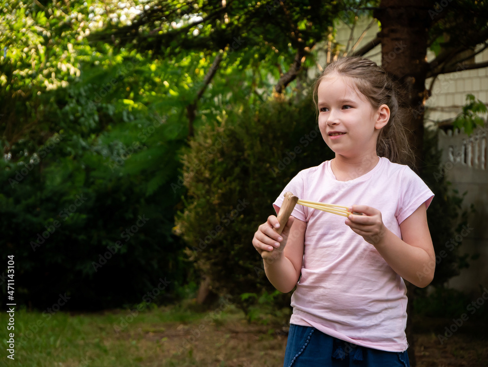 Young elementary school age child, happy cheerful girl holding a wooden toy slingshot in hands ready to shoot, outside portrait, copy space. Weapons, traditional toys natural casual outdoors scene Young elementary school age child, happy cheerful girl holding a wooden toy slingshot in hands ready to shoot, outside portrait, copy space. Weapons, traditional toys natural casual outdoors scene