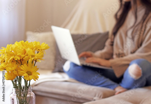 Woman types something on the computer sitting on the couch with a pot of yellow flowers by her side © leticiaalvares