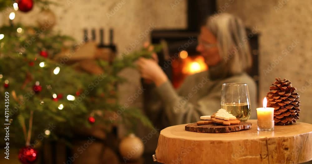 Woman decorating Christmas tree in the background. Focus on the table with a glass of wine, cookies and candlelight in front. Woman decorating Christmas tree in the background. Focus on the table with a glass of wine, cookies and candlelight in front.