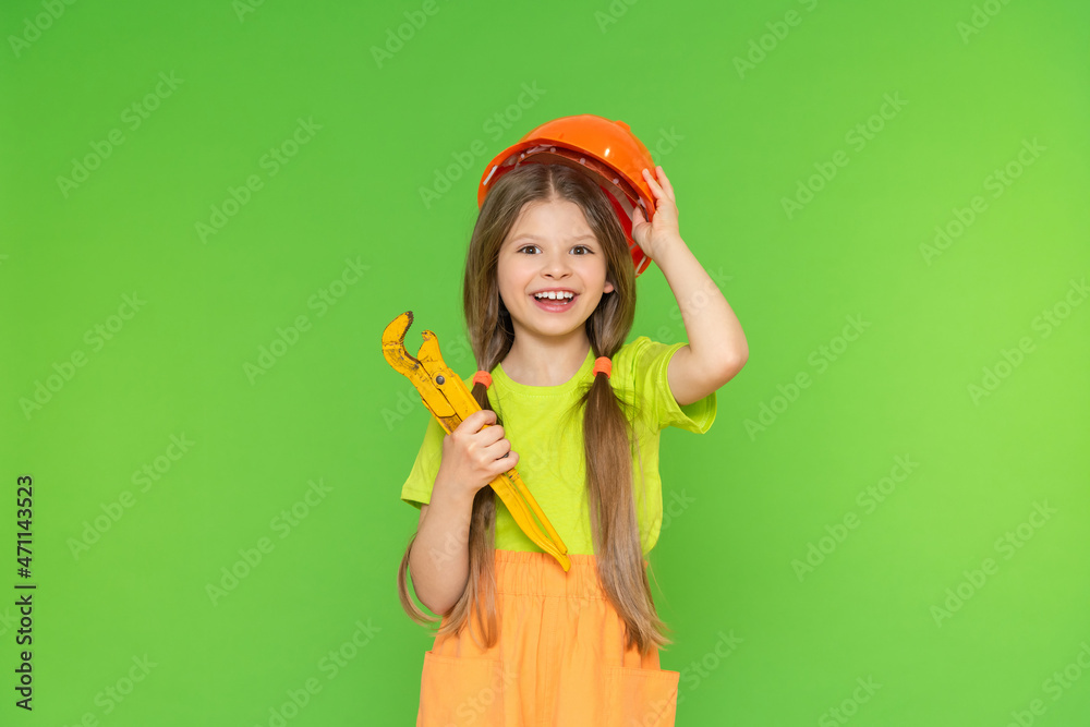 A cheerful little girl holds a construction safety helmet and a wrench. Construction and repair. Green isolated background. Copy space. A cheerful little girl holds a construction safety helmet and a wrench. Construction and repair. Green isolated background. Copy space.