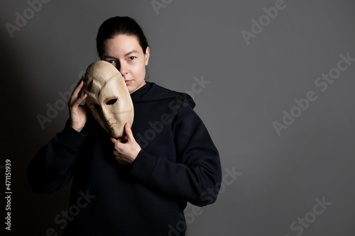 Canvas Print Hiding behind a mask, a young woman in a dark hoodie hides her face with a mask, the concept of self-doubt