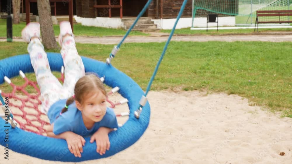 Happy cheerful elementary school age girl, child swinging on a modern round swing laying down, closeup, using playground equipment outdoors, recreation, relaxation and leisure concept, one person