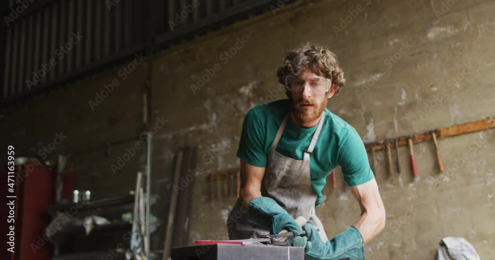 Caucasian male blacksmith wearing safety glasses, hammering hot metal tool on anvil in workshop Caucasian male blacksmith wearing safety glasses, hammering hot metal tool on anvil in workshop