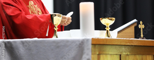 A Catholic Priest about to break the bread host while celebrating blessed Holy Communion at Mass. Wearing a red gown vestment and surrounded by chalice cross crucifix and bible photo