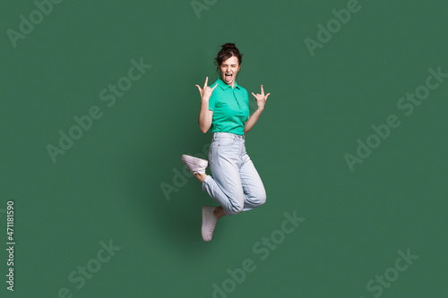 Full size photo of young crazy caucasian woman jumping showing tongue. Rock'n'roll sign. Isolated on green color background