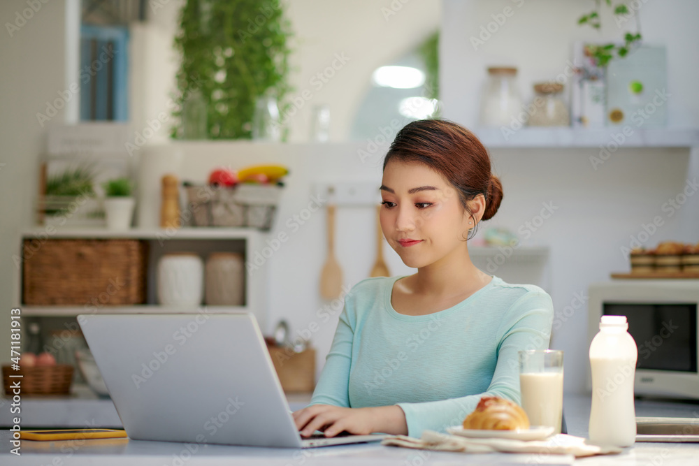 Beautiful young woman drinking healthy delicious smoothie and using laptop at home