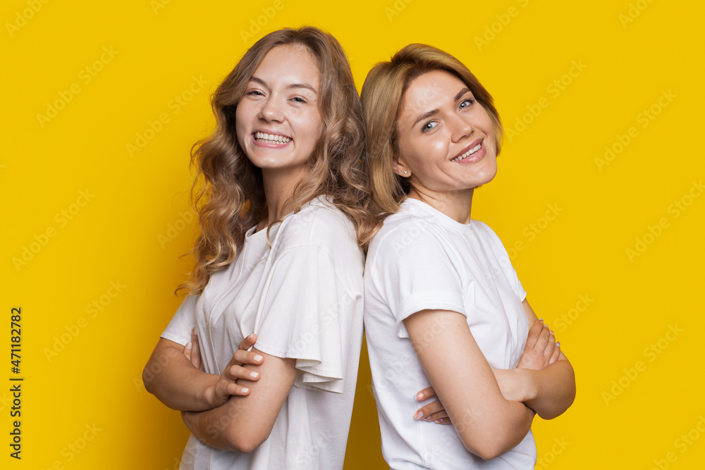 Portrait of two laughing women in white t-shirts standing back to back isolated over yellow background with arms crossed. Closeup portrait. Portrait of two laughing women in white t-shirts standing back to back isolated over yellow background with arms crossed. Closeup portrait.