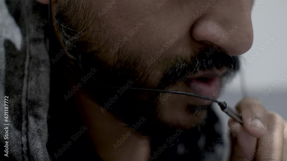 Close up shot of the face of a bearded Indian man holding a microphone from a headset in front of his mouth, talking calmly to the caller on his mobile phone