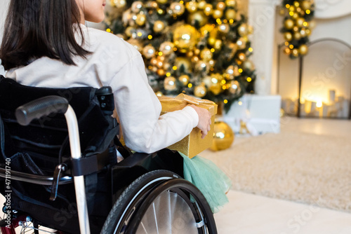 little girl in a wheelchair near the Christmas tree. disabled person holding yellow, golden New Year's gift