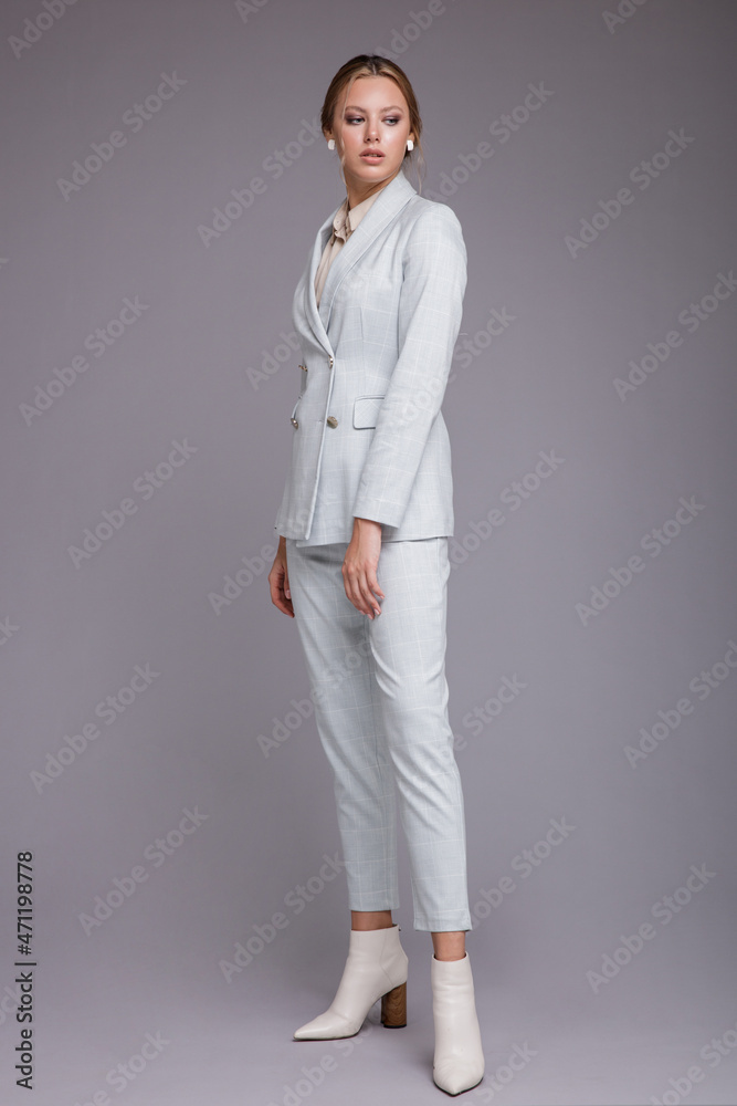 Fashion photo of a beautiful elegant young woman in a pretty soft blue suit, jacket, pants, trousers, white boots posing on gray background. Studio shot, portrait. Monochrome colors Fashion photo of a beautiful elegant young woman in a pretty soft blue suit, jacket, pants, trousers, white boots posing on gray background. Studio shot, portrait. Monochrome colors