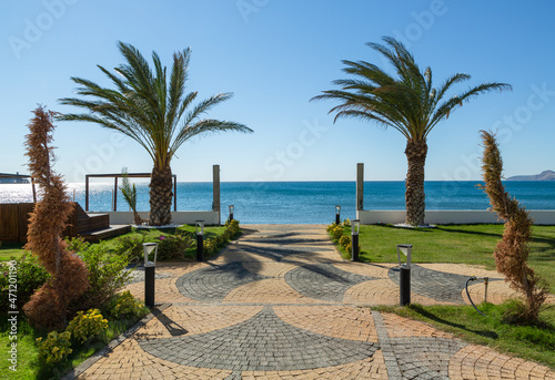 Mediterranean view through palm trees in a garden. A walking path leading down to the beach from a landscaped garden.