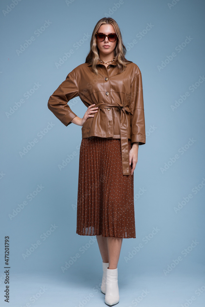 Fashion photo of a beautiful elegant young woman in a pretty brown leather jacket, brown long skirt, stylish sunglasses posing over blue background. Studio shot, portrait. Brown and blue colors Fashion photo of a beautiful elegant young woman in a pretty brown leather jacket, brown long skirt, stylish sunglasses posing over blue background. Studio shot, portrait. Brown and blue colors