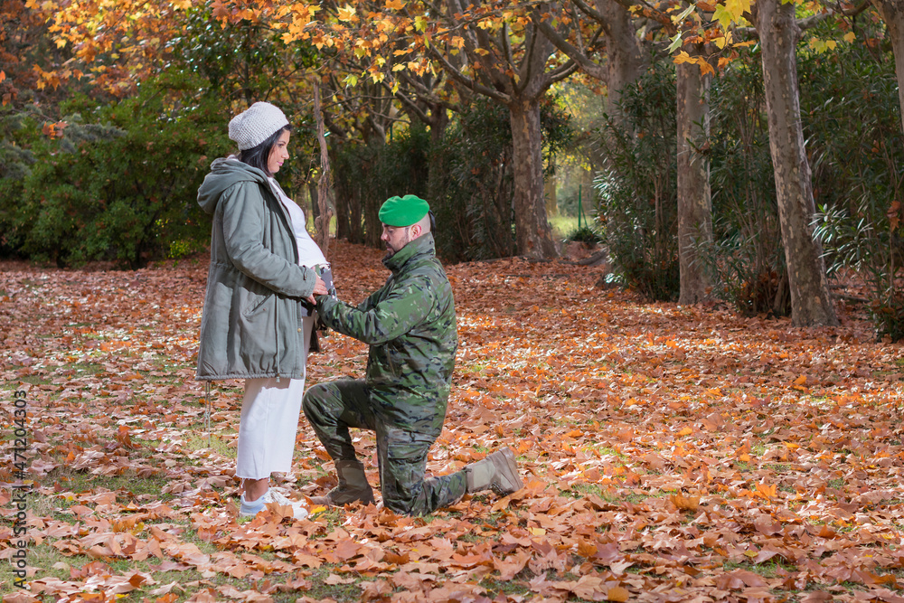Hombre militar de rodillas sostiene en el vientre de su esposa embarazada la ropa de su futuro bebé en un bosque otoñal Hombre militar de rodillas sostiene en el vientre de su esposa embarazada la ropa de su futuro bebé en un bosque otoñal